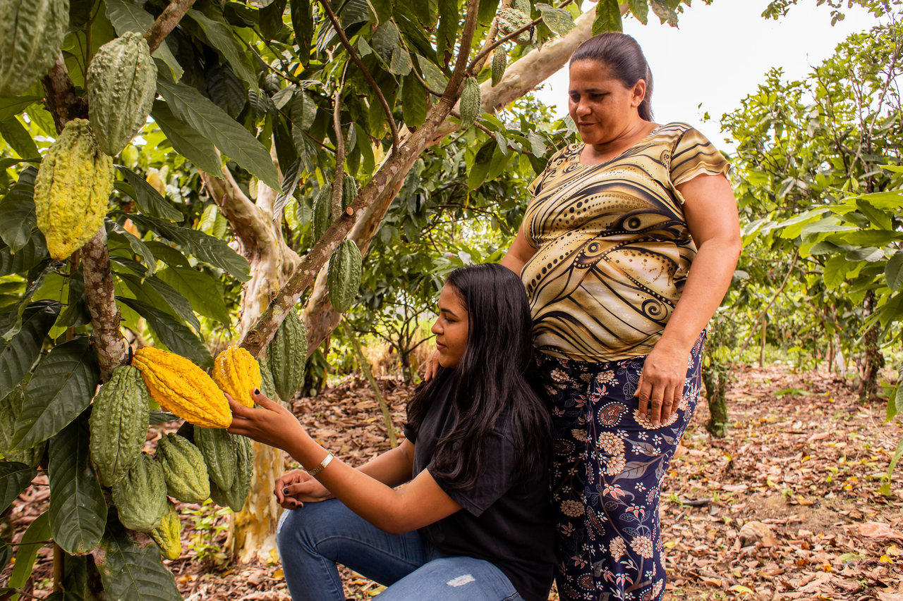 Uma adolescente segura um fruto de cacau ao lado de sua mãe em um cenário rural